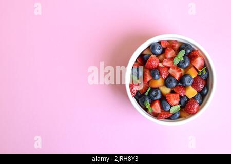 Obstsalat in einer weißen Tasse auf rosa Hintergrund. Erdbeeren, Pfirsiche und Heidelbeeren in Scheiben geschnitten mit Minzblättern. Freier Speicherplatz für Text. Stockfoto