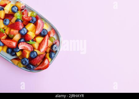 Obstsalat auf einem rechteckigen Teller auf einem rosa Hintergrund. Erdbeeren, Pfirsiche und Heidelbeeren in Scheiben geschnitten mit Minzblättern. Stockfoto
