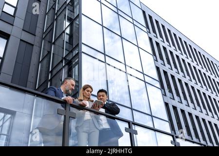 Drei Geschäftsleute, Frau und Mann, außerhalb des Bürogebäudes, ernsthaft und nachdenklich diskutieren und Blick auf elektronische Tablet, asiatische Mann und Frau in Business-Anzüge Stockfoto