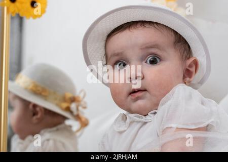 Schönes Mädchen mit einem weißen Hut, Blick erschrocken auf die Kamera. Im Hintergrund ein Spiegel mit der Reflexion des kleinen Mädchens. Stockfoto