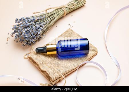 Ätherisches Lavendelöl in blauer Glasflasche mit Pipette. Flasche mit duftendem Öl und Lavendelblüten auf beigem Hintergrund. Nachbau von Lavendel essent Stockfoto