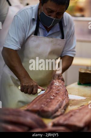 Ein Metzger trägt eine Maske und schneidet ein großes Stück Fleisch in einem Laden Stockfoto