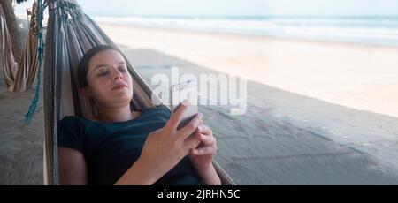 Schöne junge hispanische Frau in einem blauen T-Shirt, die sich in einer Hängematte ausruht und an einem sonnigen Tag Selfies mit ihrem Smartphone am Strand macht Stockfoto