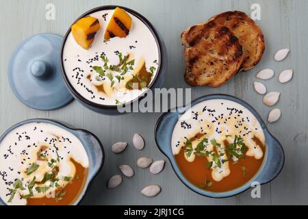 Cremige Kürbissuppe mit Rahm, schwarzen Kreuzkümmelkernen, Kräutern, Kürbiskernen und geröstetem Brot auf einem blauen Holztisch Stockfoto