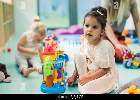 Spielerisch im Kindergarten lernen. Kleinkind kleines Mädchen und die Lehrerin spielen mit bunten Kunststoff-Spielhäusern, Bausteinen, Autos und Booten. Entwicklung von Phantasie, Kreativität, Feinmotorik und grobmotorischen Fähigkeiten. Hochwertige Fotos Stockfoto