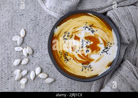 Eine Platte aus cremiger Kürbissuppe mit cremefarbenem und schwarzem Kreuzkümmel und Kürbiskernen auf einem leichten Strickpullover aus Wolle Stockfoto