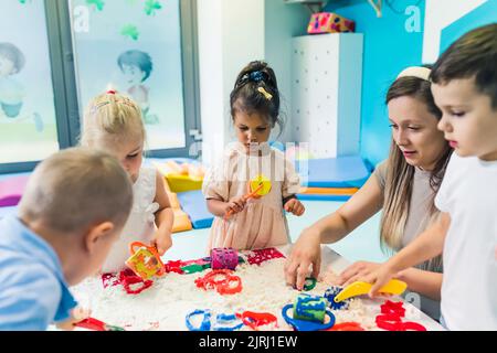 Sensorisches Spielen in der Kinderschule. Kleinkinder mit ihrem Lehrer haben Spaß mit weißen, flauschigen Wolkenteig und verwenden Modellierwerkzeuge wie bunte und strukturierte Rollstifte, Schneider, Silikon-Formkoffer. Kreativität und Vorstellungskraft Entwicklung . Hochwertige Fotos Stockfoto