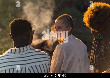 Eine multikulturelle Gruppe von Freunden, die in der Natur am Lagerfeuer angenehme Sonnenuntergänge genießen, während sie auf dem Gras sitzen und miteinander reden. Hochwertige Fotos Stockfoto