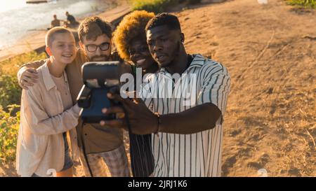 Multiethnische Gruppe von besten Freunden, die Erinnerungen schaffen, indem sie lustige Fotos mit einer alten polaroid-Kamera machen und sich in der Nähe des Flusses bei Sonnenuntergang im Sommer entspannen. Hochwertige Fotos Stockfoto