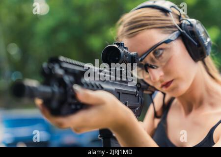 Nahaufnahme einer Lupe eines schwarzen Gewehr, das von einer fokussierten, mächtigen kaukasischen Frau in schützendem Schießstand gehalten wird. Unscharfer Hintergrund. Hochwertige Fotos Stockfoto