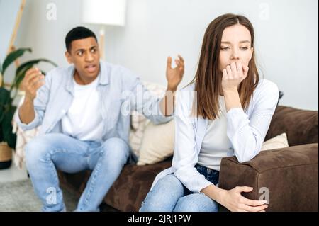 Gemischtes Rennpaar sitzt auf einem Sofa im Wohnzimmer, ein wütender Mann schreit eine Frau an, fühlt sich wütend, frustriert, traurig Mädchen von ihm abgewandt. Missverständnisse in Beziehungen, Eifersucht, Streit Stockfoto