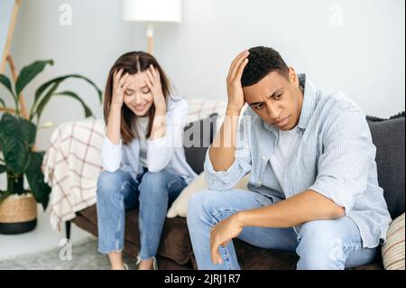 Streit, Groll gegeneinander. Verärgerte traurige Ehegatten, ein multirassisches Paar, sitzen auf einem Sofa im Wohnzimmer, beleidigt von einander nach einem Showdown und Schreien Stockfoto
