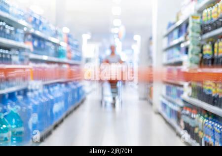 Verschwommener Supermarkt Gang mit Regalen von Waren. Perspektivische Ansicht des abstrakten Lebensmittelgeschäftes Gang - Frau Kundin mit Warenkorb in der Mitte Stockfoto