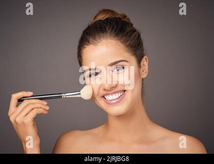 Bürsten auf rosigen Wangen. Porträt eines schönen jungen Models, das im Studio ihre Wangen mit einem Blusher befeuert. Stockfoto