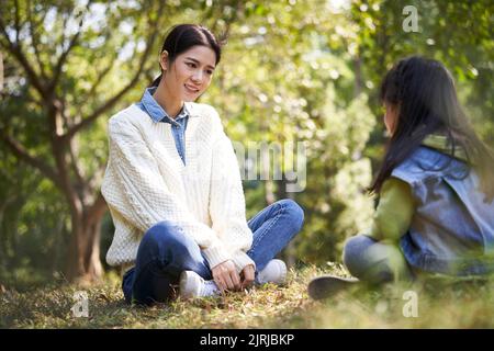 Junge asiatische Mutter sitzt auf Gras im Park mit einem angenehmen Gespräch mit Vorschultochter Stockfoto