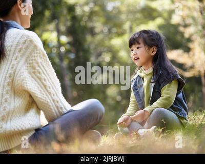 Junge asiatische Mutter sitzt auf Gras im Park mit einem angenehmen Gespräch mit Vorschultochter Stockfoto