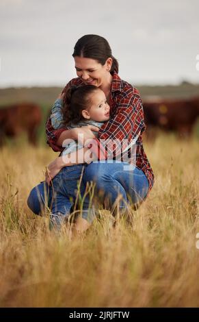 Liebe, Familie und Fürsorge mit einer Mutter und Tochter, die sich auf einem Feld draußen auf einem Bauernhof umarmen. Viehzüchter und kleines Mädchen in der Landwirtschaft, Landwirtschaft und Stockfoto