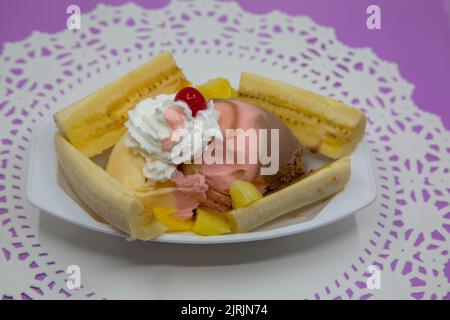 Bananeneis-Eisbecher auf weißer Deckchen und violetter Tischplatte. Neapolitanisches Eis mit Schlagsahne, Bananen, Maraschino-Kirsche und Ananas Stockfoto