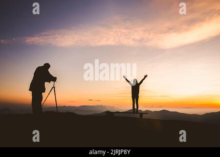 Silhouette des Fotografen, der bei Sonnenuntergang mit einem Model auf dem Berg fotografiert, professionelle Hochzeitsfotografie Stockfoto