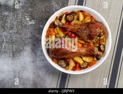 Gebackene putenbeine mit Gemüse - Kartoffeln, Auberginen, Karotten und Paprika in einer weißen Backform auf dunkelgrauem Hintergrund. Draufsicht, flach liegend Stockfoto
