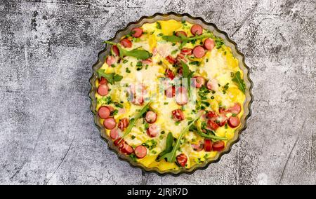 Wurst Frittata mit Käse, Rucola, Kartoffeln in einer Glasbackform auf dunkelgrauem Hintergrund. Draufsicht, flach liegend Stockfoto