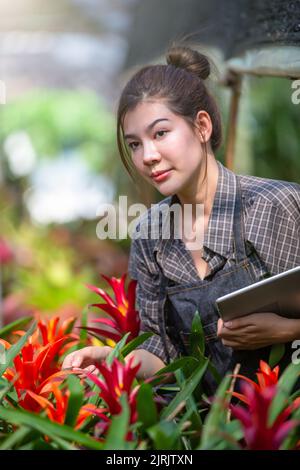 Ein schöner Bauer arbeitet in einem Gewächshaus an Blumen und notiert sich auf einer Tablette. Frühling und Sommer. Geschäftskonzept mit kleinen Blumenmustern. Stockfoto