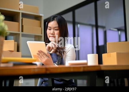 Charmante und talentierte junge asiatische weibliche Online-Shop-Besitzerin, die ihren Lagerbestand überprüft und ihren Produktstatus auf der Website auf einem digitalen Tablet aktualisiert. KMU-Bus Stockfoto