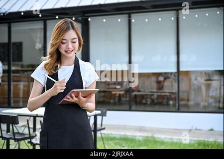 Attraktive und schöne junge asiatische Barista oder Kellnerin in ihrer Uniform, die mit ihrem digitalen Tablet vor dem Café steht. Stockfoto