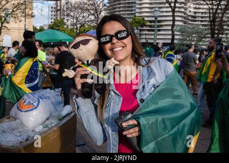 Belo Horizonte, Brasilien. 24. August 2022. Ein Anhänger des brasilianischen Präsidenten Jair Bolsonaro trägt eine Sonnenscheibe mit Bolsonaros Namen auf seinen Kristallen und zeigt eine Puppe des rechtsextremen Kandidaten während einer Wahlkampfveranstaltung im Rahmen seines Wiederwahlkampfs im Praça da Liberdade in Belo Horizonte, im brasilianischen Bundesstaat Minas Gerais. Kredit: SOPA Images Limited/Alamy Live Nachrichten Stockfoto