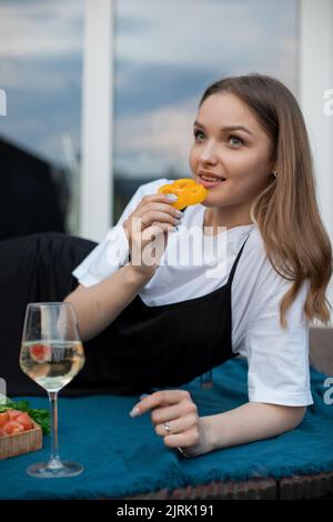 Porträt einer jungen Frau, die auf einer blauen Decke in der Nähe eines Glases Weißwein liegt, Früchte auf einem Holztablett und ein Stück Pfeffer isst. Stockfoto