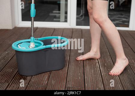 Beschnittenes Foto einer jungen Barfußfrau, die einen Spinnmopp über einen grauen Plastikeimer hält und den Holzboden der Veranda reinigt. Stockfoto