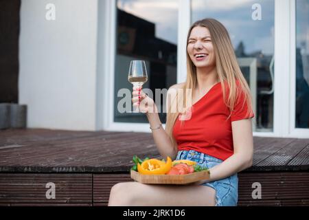 Porträt einer jungen lachenden hübschen Frau, die auf einer hölzernen Veranda sitzt, ein Glas Weißwein hält und Gemüse schneidet. Stockfoto