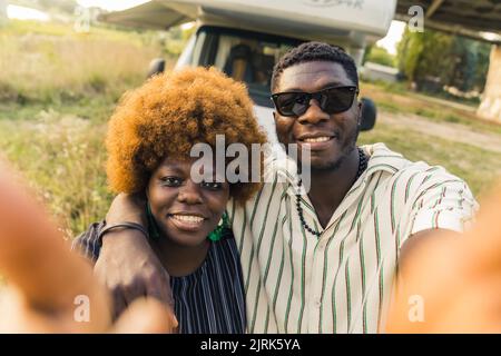 Lächelndes, ethnisches, dunkelhäutig gerades Paar, das im Sommer einen Roadtrip in einem Campingbus macht, sich umarmt und ein Selfie vor ihrem Fahrzeug macht. Hochwertige Fotos Stockfoto