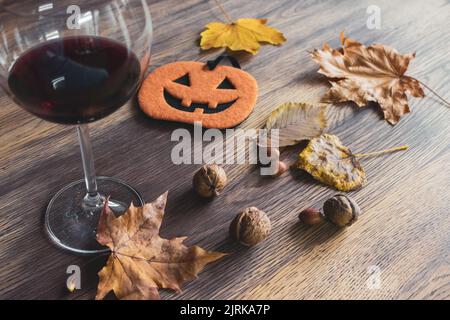 Halloween Stillleben. Ein Glas Rotwein, Nüsse, trockene Blätter und lustige Kürbisfigur auf dem Tisch. Herbstferien. Oktober Hintergrund. Stockfoto