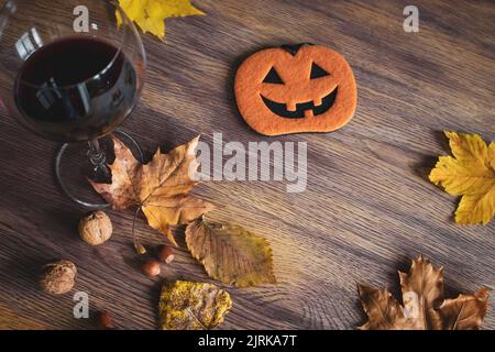 Halloween Stillleben. Ein Glas Rotwein, Nüsse, trockene Blätter und lustige Kürbisfigur auf dem Tisch. Herbstferien. Oktober Hintergrund. Stockfoto