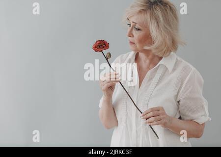 Traurige ältere Frau im weißen Hemd hält eine trockene, alte, tote Rosenblume. Alter und Alterungskonzept. Stockfoto