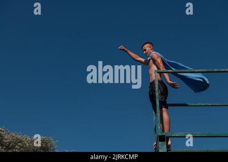 Kleiner Teenager-Junge am Fluss, der sich mit Freunden auf einer Sommerparty amüsieren kann und ein Handtuch als Superhelden-Schal trägt, der bereit für einen Sprung ist, der mutig in der Ferne aussieht Stockfoto