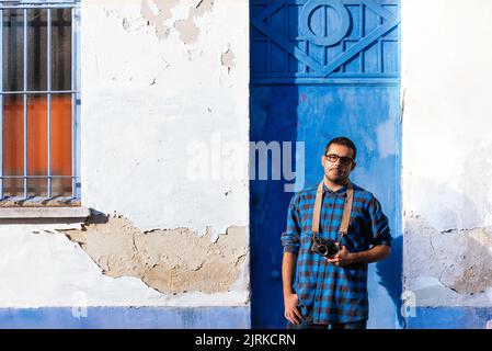 Junger charismatischer männlicher Fotograf in legerer Kleidung, der in der Nähe des schäbigen Gebäudes steht und die Kamera anschaut Stockfoto