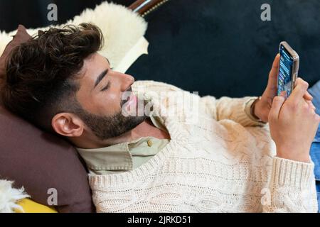 Von oben auf Inhalt junge unrasierte ethnische Männer in stilvollen Strickpullover liegend auf dem Sofa und lächelnd, während Messaging auf dem Smartphone zu Hause Stockfoto