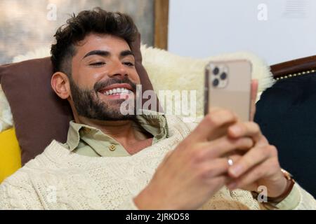 Von oben auf Inhalt junge unrasierte ethnische Männer in stilvollen Strickpullover liegend auf dem Sofa und lächelnd, während Messaging auf dem Smartphone zu Hause Stockfoto