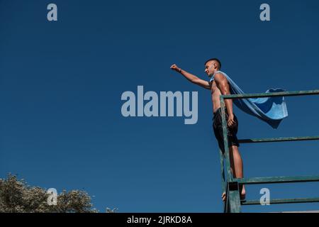 Kleiner Teenager-Junge am Fluss, der sich mit Freunden auf einer Sommerparty amüsieren kann und ein Handtuch als Superhelden-Schal trägt, der bereit für einen Sprung ist, der mutig in der Ferne aussieht Stockfoto