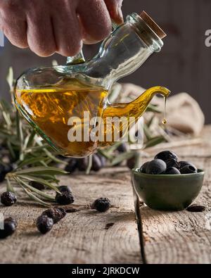 Unkenntlich: Der Koch gießt frisches Öl auf reife schwarze Oliven über den Holztisch in der Nähe von Ästen Stockfoto