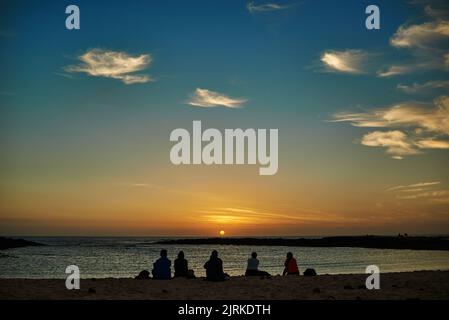 Rückansicht anonymer Silhouetten von Reisenden, die beim Sonnenuntergang auf Fuerteventura Spanien das wellige Meer unter bewölktem Himmel betrachten Stockfoto