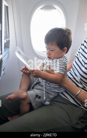 Kleinkind, das im Flugzeug auf dem MutterSchoß sitzt. Ein Jahr alter Junge, der im Flugzeug fliegt und versucht, sich zu unterhalten Stockfoto