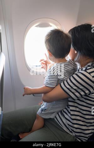 Das Kleinkind, das im Flugzeug sitzt und auf seinem MutterSchoß sitzt, blickt beide aus einem Flugzeugfenster. Ein Jahr alter Junge, der im Flugzeug fliegt und versucht zu fliegen Stockfoto