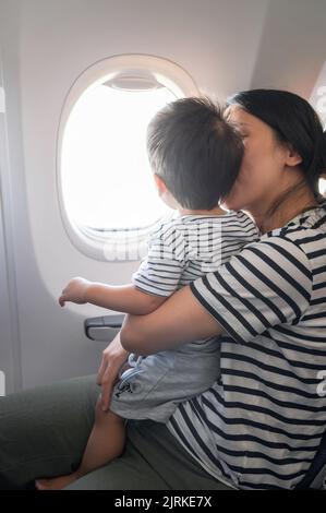 Das Kleinkind, das im Flugzeug sitzt und auf seinem MutterSchoß sitzt, blickt beide aus einem Flugzeugfenster. Ein Jahr alter Junge, der im Flugzeug fliegt und versucht zu fliegen Stockfoto