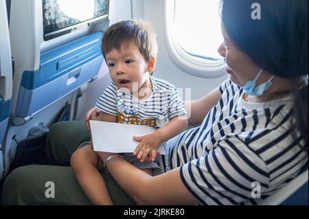Kleinkind, das im Flugzeug auf dem MutterSchoß sitzt. Ein Jahr alter Junge, der im Flugzeug fliegt und versucht, sich zu unterhalten Stockfoto