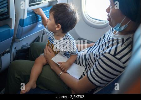 Kleinkind, das im Flugzeug sitzt, auf seinem MutterSchoß und den Bildschirm des Flugunterhaltungssitzes benutzt. Ein Jahr alter Junge, der im Flugzeug fliegt und versucht zu en Stockfoto
