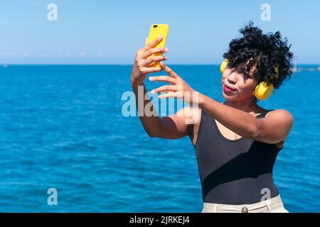 Afroamerikanische Frau mit lockigen Haaren in kabellosen Kopfhörern, die sich auf dem Handy gegen das Meer im Sonnenlicht porträtiert Stockfoto