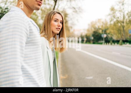 Seitenansicht des Inhalts stilvolle junge Frau, die Hände eines beschnittenen, nicht erkennbaren Freundes hält, während sie die Straße überquert, in der Stadt spazieren geht und wegschaut Stockfoto